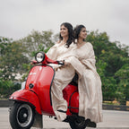 Two women in white sarees sitting on a red scooter with a blurred background